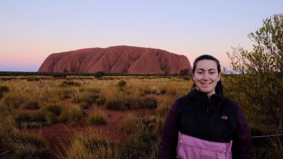 Sheila stands in front of Uluru at Uluru-Kata Tjuta National Park in the Northern Territory, which is a sacred spot to the aboriginal people of Australia. She is there to watch the sunset and watch the rock's color shades change as the sun light hits it