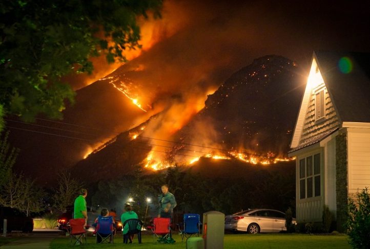 A fire burns a mountainside above a peaceful scene of people sitting in folding chairs outside a nighttime church