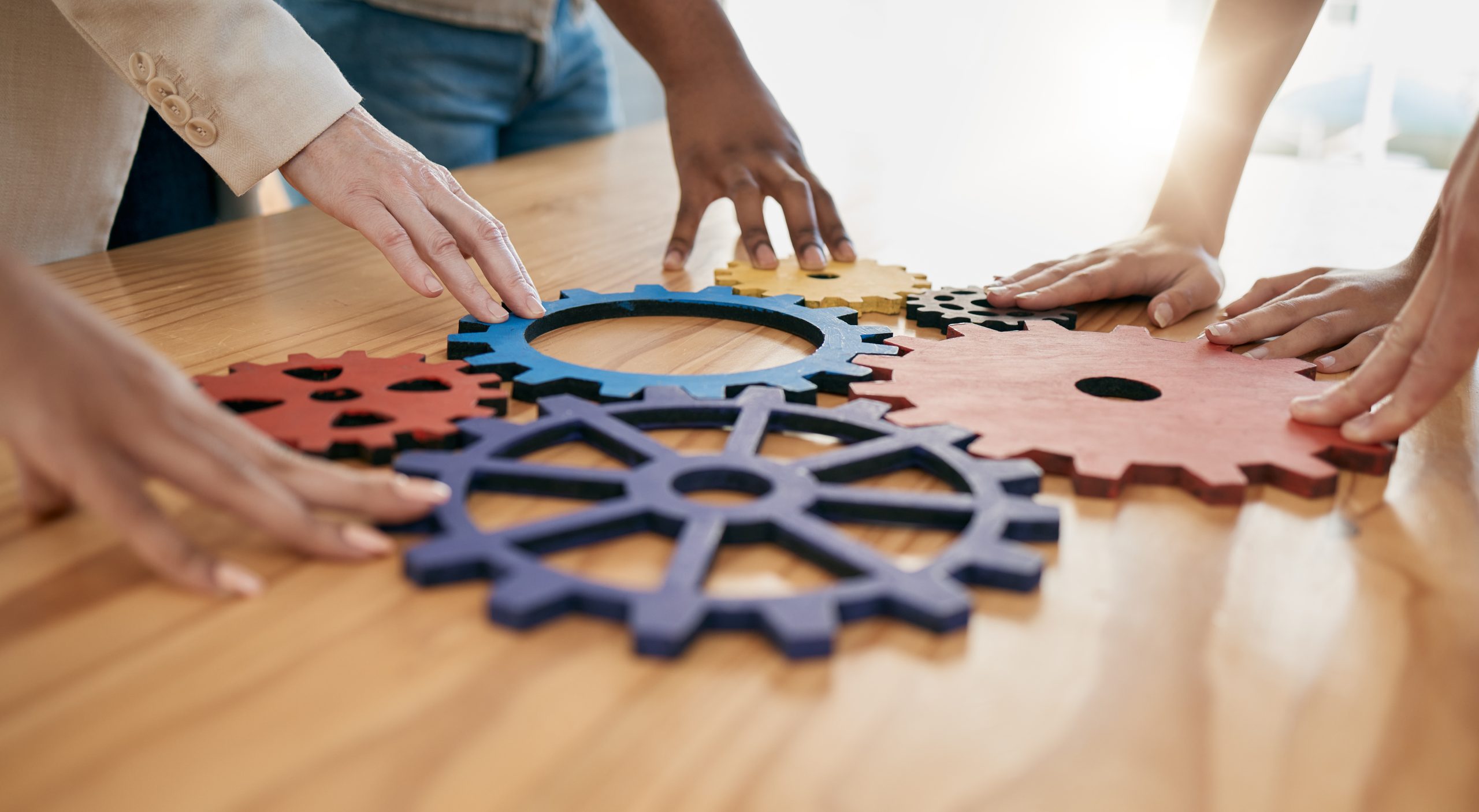 A table with symbolic wooden gears on it, with multiple peope's hands touching the gears.